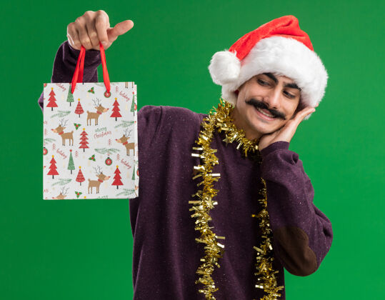 举行快乐的小胡子男人戴着圣诞老人的帽子 脖子上挂着金属丝 手里拿着圣诞礼物的纸袋 站在绿色的背景上 面带微笑地看着它年轻帽子圣诞节