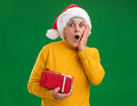 盒子震惊的老妇人戴着圣诞老人的帽子 把手放在脸上 拿着一个紫色背景的圣诞礼盒 里面有复制空间举行手老人