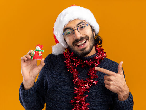 点微笑的年轻帅哥戴着圣诞帽 脖子上戴着花环 指着橙色背景上孤立的玩具花环圣诞节家伙