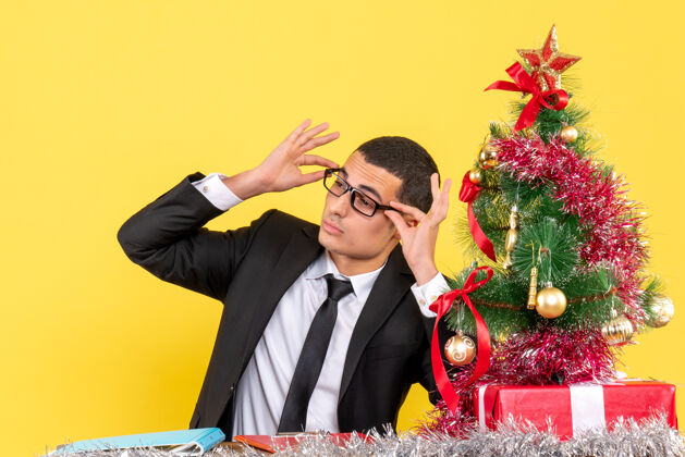 礼物前视图年轻人坐在桌边摘下眼镜圣诞树和礼物圣诞树专业年轻