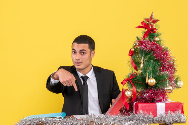 坐着正面图西装革履的男人坐在桌子旁拿着文件 手指着圣诞树和礼物圣诞树西装革履的男人拿着