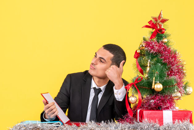 做正面图西装革履的男人坐在桌子旁看着右手竖起大拇指的牌子圣诞树和礼物人树起来