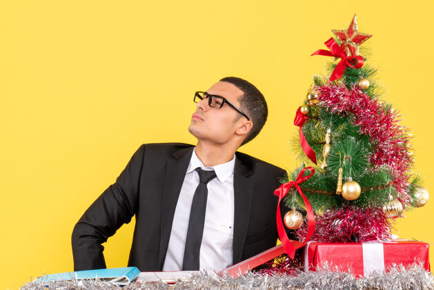 圣诞节前视图戴眼镜的年轻人坐在桌子旁看着左边的圣诞树和礼物左边专业人士坐着