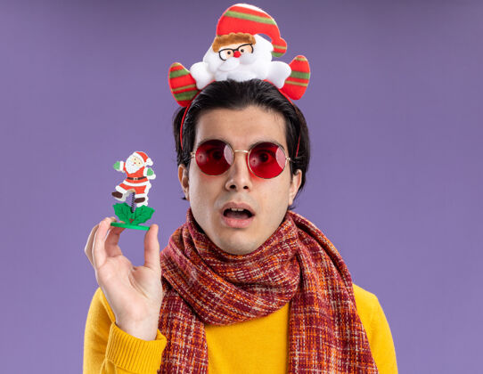 惊讶脖子上围着暖围巾的年轻人 穿着黄色高领毛衣 头上戴着滑稽的镶边 手里拿着圣诞玩具 惊讶地站在紫色的墙上高领毛衣轮辋脖子