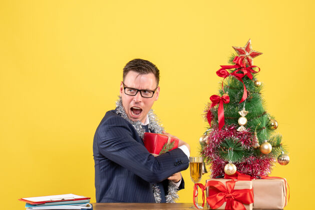 前面焦躁不安的男人把礼物藏在圣诞树旁的桌子上 黄色的墙上挂着礼物树成人圣诞节