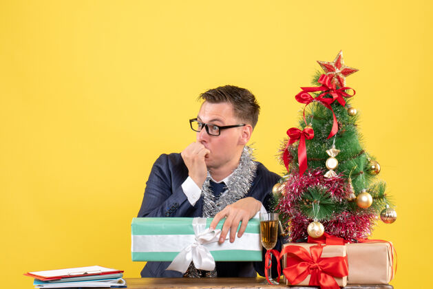 圣诞节一个好奇的男人坐在圣诞树旁的桌子前看着什么东西 黄色的礼物公司奇怪的礼物