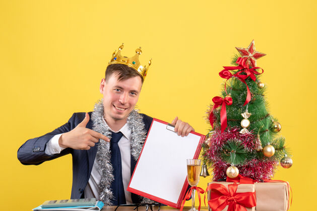 快乐前视图的快乐男子指着剪贴板坐在桌旁圣诞树和黄色的礼物商人新郎人