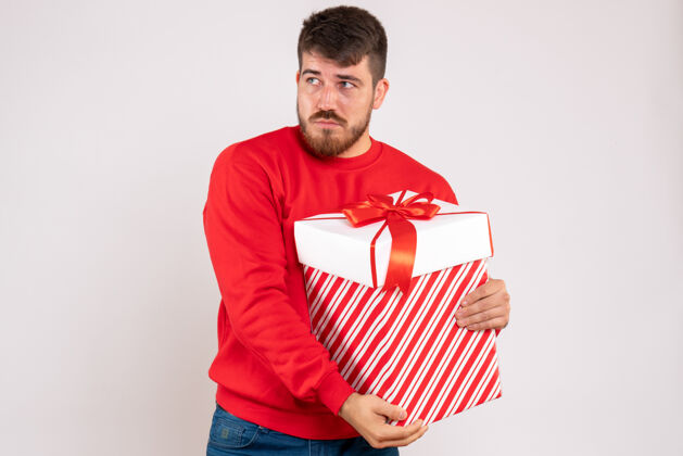 抱着前视图中的年轻人在红衫举行圣诞礼物在一个白色的墙壁框成人圣诞节年轻