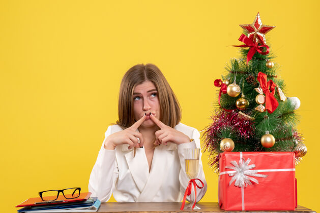 快乐正面图：女医生坐在黄色背景的桌子前 放着圣诞树和礼品盒女医生圣诞节漂亮