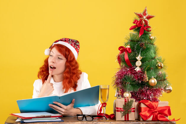 电脑正面图：女医生坐在桌子后面看黄色背景上的文件 还有圣诞树和礼品盒圣诞节办公室工作