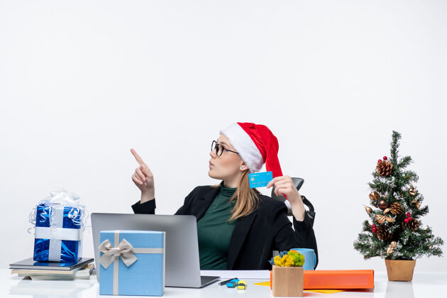女人戴着圣诞老人帽戴着眼镜的金发女人坐在桌子旁 手里拿着圣诞礼物和银行卡摩托宝抱着金发