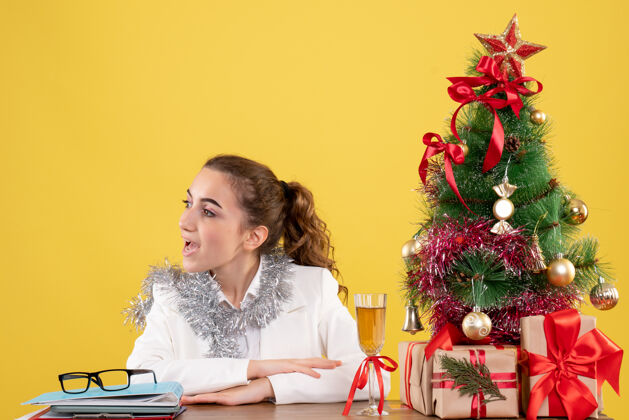 护士正面图：女医生坐在黄色背景的桌子后面 还有圣诞树和礼品盒成人微笑办公室