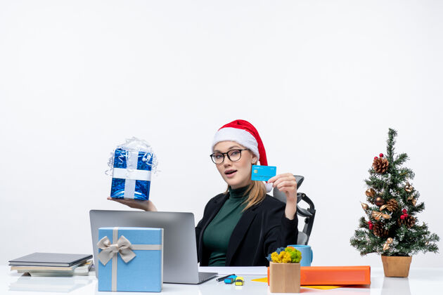 卡片一个戴着圣诞老人帽子戴着眼镜的女商人坐在桌子旁 手里拿着圣诞礼物和银行卡自由职业者圣诞老人圣诞树