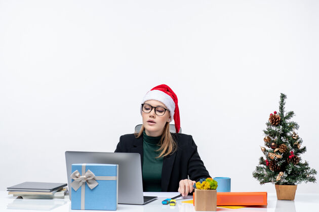 笔记本电脑戴着圣诞老人帽的女商人坐在桌子旁 桌上放着圣诞树和礼物女士工作圣诞树
