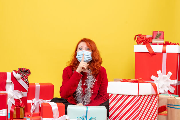 圣诞节一个戴着面具坐在黄色墙壁上的年轻女子围着礼物的正视图病毒Covid成人