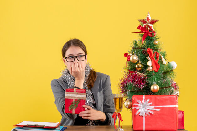 人前视图困惑的女孩戴着眼镜坐在桌子旁看着圣诞树和礼物鸡尾酒圣诞节坐着女孩