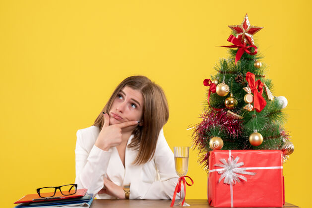 健康正面图：女医生坐在黄色背景的桌子前 放着圣诞树和礼品盒情绪圣诞节前面