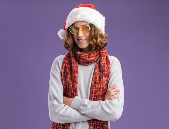 站着一个戴着圣诞老人帽 戴着黄色眼镜 脖子上围着暖和围巾的年轻人看着镜头 自信地微笑着 双臂交叉着站在紫色的背景上相机脖子圣诞老人