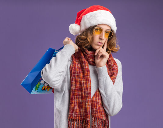 相机戴着圣诞老人帽 戴着黄色眼镜 脖子上围着暖和围巾的年轻人手里拿着圣诞纸袋 手里拿着礼物 严肃地看着站在紫色背景下的相机严肃看年轻