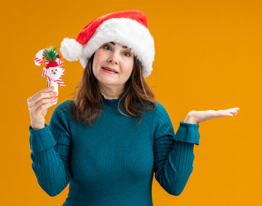成人惊讶的成年白人妇女戴着圣诞帽 手持糖果手杖 在橙色背景上保持手张开 并留有复制空间手杖打开快乐