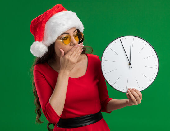 女孩戴着圣诞帽 戴着眼镜 手放在嘴上 看着时钟 在绿色背景下与世隔绝的年轻漂亮女孩关心帽子嘴巴