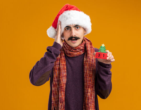 帽子一个留着胡子的男人戴着圣诞老人帽 脖子上围着暖和的围巾 手里拿着新年礼物的玩具立方体 站在橙色背景下焦急地看着相机橙色围巾温暖