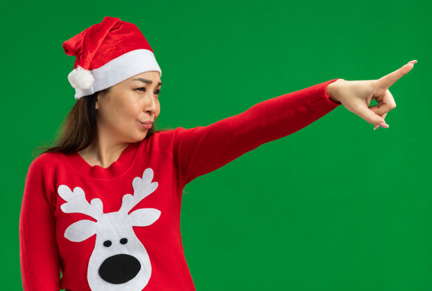 一边戴着圣诞老人帽 穿着红色毛衣的年轻女子 严肃的面带微笑 用食指指着绿色的背景圣诞节毛衣认真的
