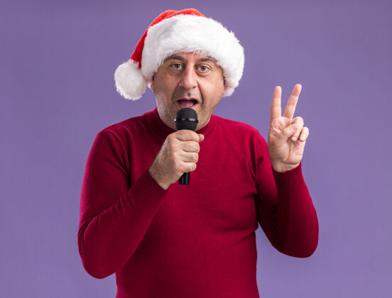 年龄快乐的中年男人戴着圣诞老人的帽子对着麦克风说话展示着v型标志看着紫色背景下的摄像机站着说话圣诞节