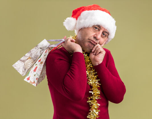 中间中年男子戴着圣诞老人帽子 脖子上戴着金属丝 手里拿着装有圣诞礼物的纸袋 站在绿色的背景下 看着相机 又累又无聊累了脖子站着