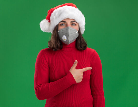圣诞老人穿着红色毛衣 戴着圣诞老人帽的年轻女孩 戴着护面面具 严肃的脸 用食指指着站在绿色墙上的一边手指严肃防护