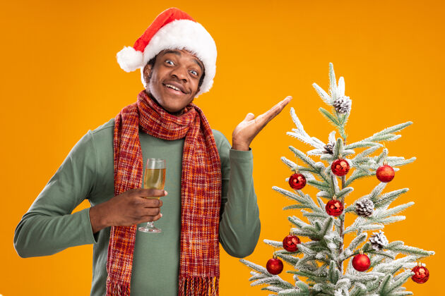 围着戴着圣诞帽 脖子上围着围巾 手举香槟的非裔美国人站在橙色墙上的圣诞树旁 愉快地微笑着男人脖子圣诞老人