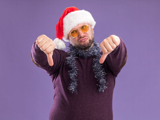 帽子一个戴着圣诞老人帽 脖子上戴着金箔花环 戴着眼镜的中年男子 一边看着紫色的墙上竖着大拇指圣诞节拇指金属片
