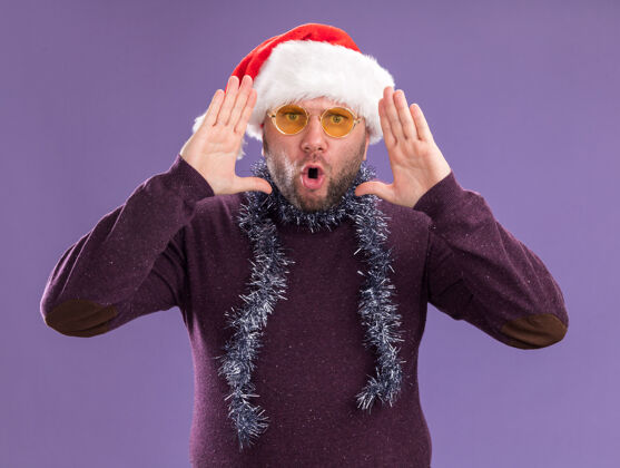 圣诞老人令人印象深刻的中年男子戴着圣诞老人帽 脖子上戴着金箔花环 戴着眼镜 手靠近头部 在紫色的墙上与世隔绝脖子中年头