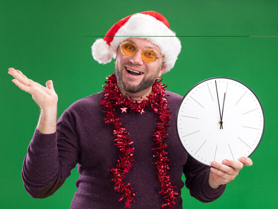 空快乐的中年男子戴着圣诞帽 脖子上戴着金箔花环 戴着眼镜 手里拿着一只被隔离在绿墙上的空指针脖子穿着金属丝