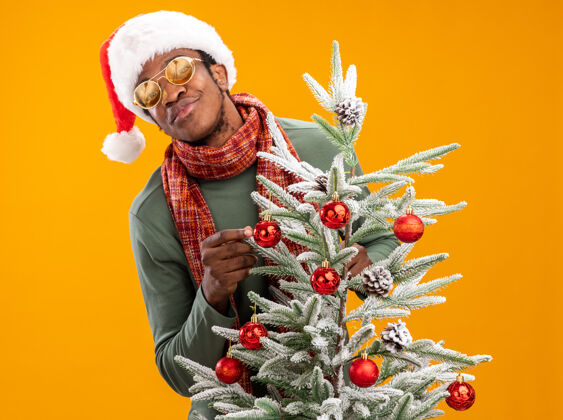 非洲人戴着圣诞帽 脖子上围着围巾 脸上带着微笑的非裔美国人站在橙色墙上的圣诞树旁脖子男人帽子
