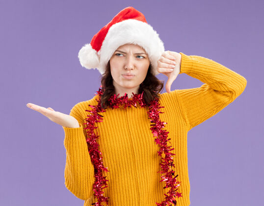 斯拉夫戴着圣诞帽 脖子上戴着花环 拇指朝下 手一直开着 在紫色背景上 留着复制空间 这是一个未被释放的年轻斯拉夫女孩周围年轻手