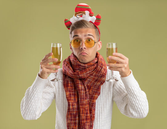 不知所措笨手笨脚的年轻帅哥戴着圣诞老人的头带和围巾拿着两杯香槟隔离在橄榄绿的墙上圣诞节围巾圣诞老人