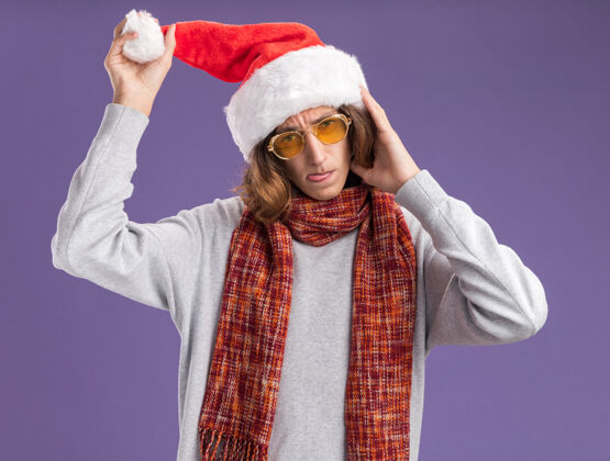 眼镜戴着圣诞老人帽 戴着黄色眼镜 脖子上围着暖和的围巾的年轻人 摸着帽子 露出舌头站在紫色的背景上 显得既高兴又高兴坚持感动周围