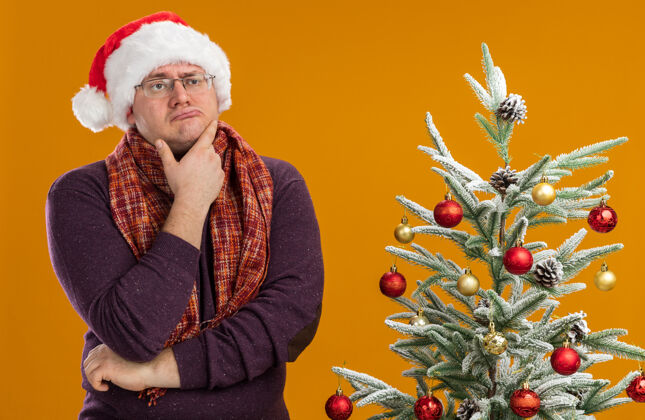橙色可疑的成年男子戴着眼镜 戴着圣诞帽 脖子上围着围巾 站在装饰好的圣诞树旁 手放在下巴上 看着橙色背景上孤立的一面圣诞快乐保持眼镜
