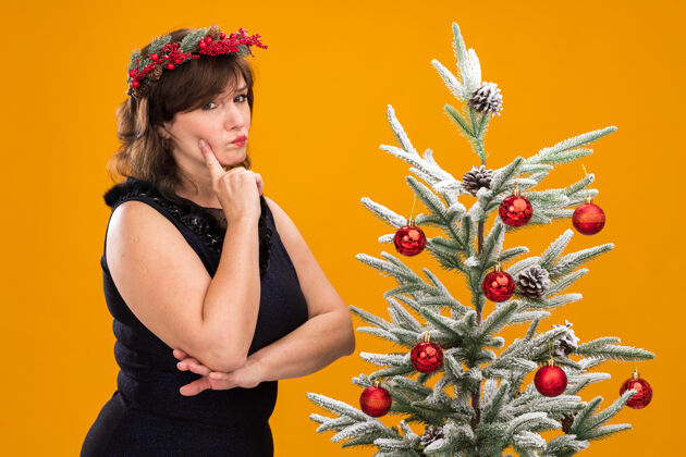 橙色可疑的中年妇女头戴圣诞花环 脖子上戴着金属丝花环 站在装饰过的圣诞树旁 在侧视图中抚摸着她的脸圣诞快乐圣诞轮廓