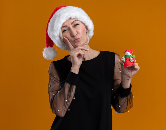 手体贴的中年金发女人戴着圣诞帽 手里拿着小雪人雕像 看着相机 手放在下巴上 在橙色背景下隔离开来金发体贴圣诞节