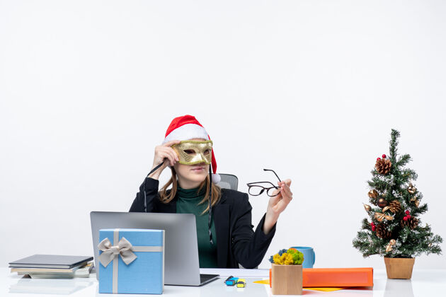 帽子戴着圣诞老人帽 戴着眼镜 戴着面具的女商人坐在桌子旁 手里拿着圣诞树和礼物树笔记本面具