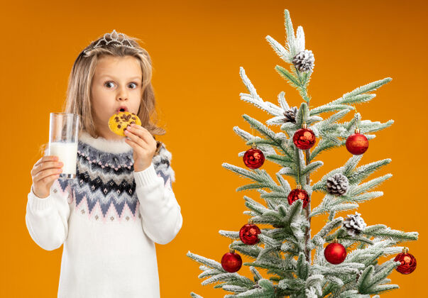 花环惊讶的小女孩站在圣诞树旁 戴着头饰 脖子上戴着花环 手里拿着一杯牛奶 尝试着橙色背景下的饼干饼干圣诞节手捧