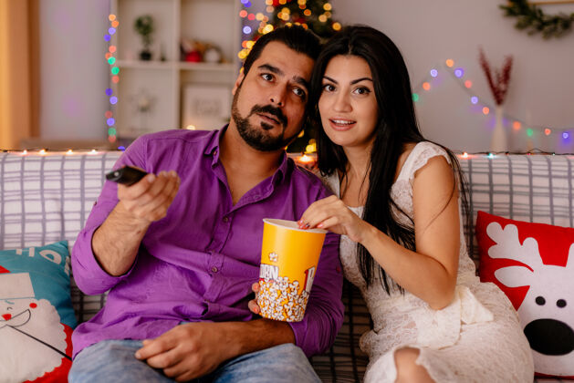 桶年轻漂亮的夫妇幸福的女人和男人坐在沙发上 拿着一桶爆米花 在装饰好的房间里一起看电视 背景是圣诞树男人树年轻