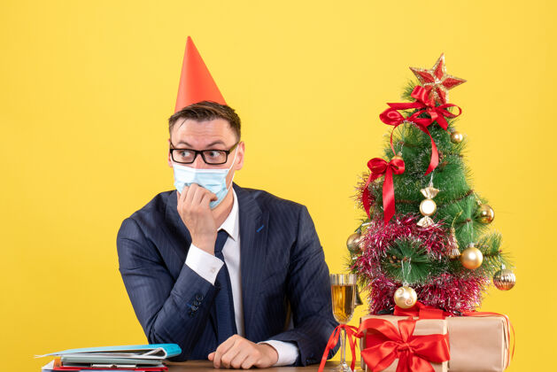 生意人戴着面具和派对帽的商人坐在圣诞树旁的桌子前 黄色的礼物男人生意男人