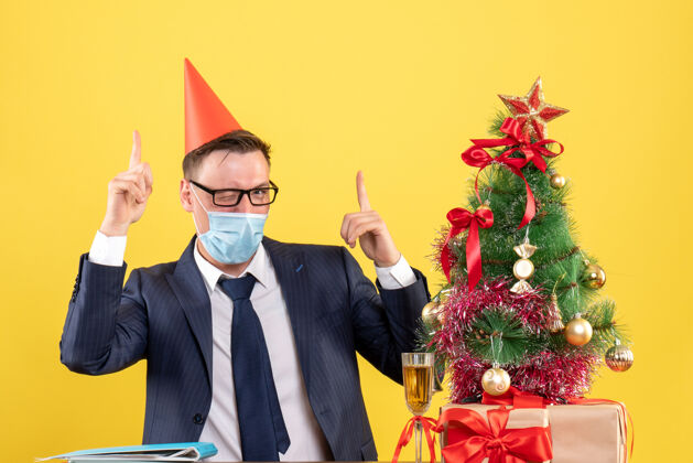 眼睛前视图中的商人戴着医用面罩眨着眼睛坐在圣诞树旁的桌子上 呈现黄色树坐着圣诞节
