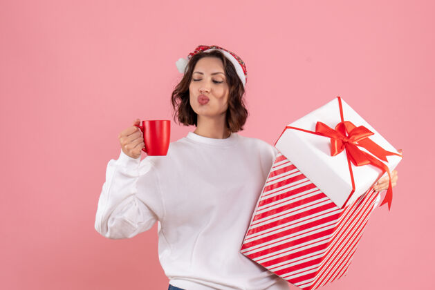 成人年轻女子手持圣诞礼物和粉红色墙上的一杯茶的前视图年轻前面年轻女性