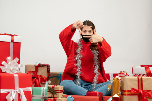 坐着穿着红色毛衣 戴着黑色面具的年轻女孩正在制作相机标志 围坐在白色的礼物上毛衣圣诞节年轻