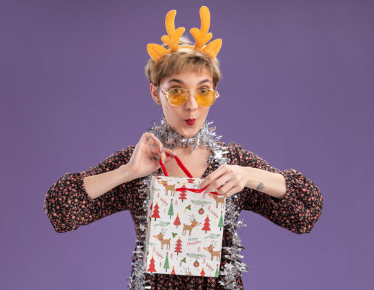 开放好奇的年轻漂亮女孩戴着驯鹿鹿角头带 脖子上戴着金箔花环 戴着眼镜 手里拿着圣诞礼品袋 把它孤立地打开在紫色的墙上好奇年轻周围