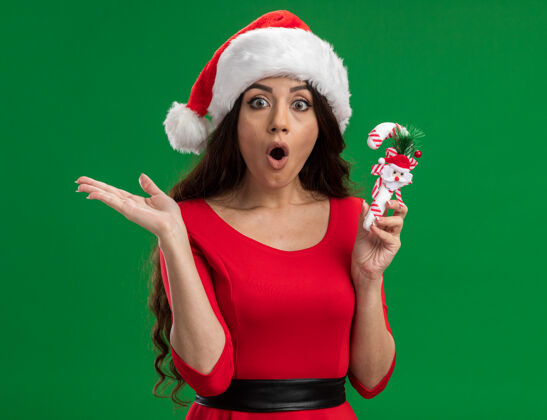 糖果惊讶的年轻漂亮女孩戴着圣诞帽拿着糖果手杖装饰看着相机显示空手孤立在绿色的背景拿着圣诞老人惊讶
